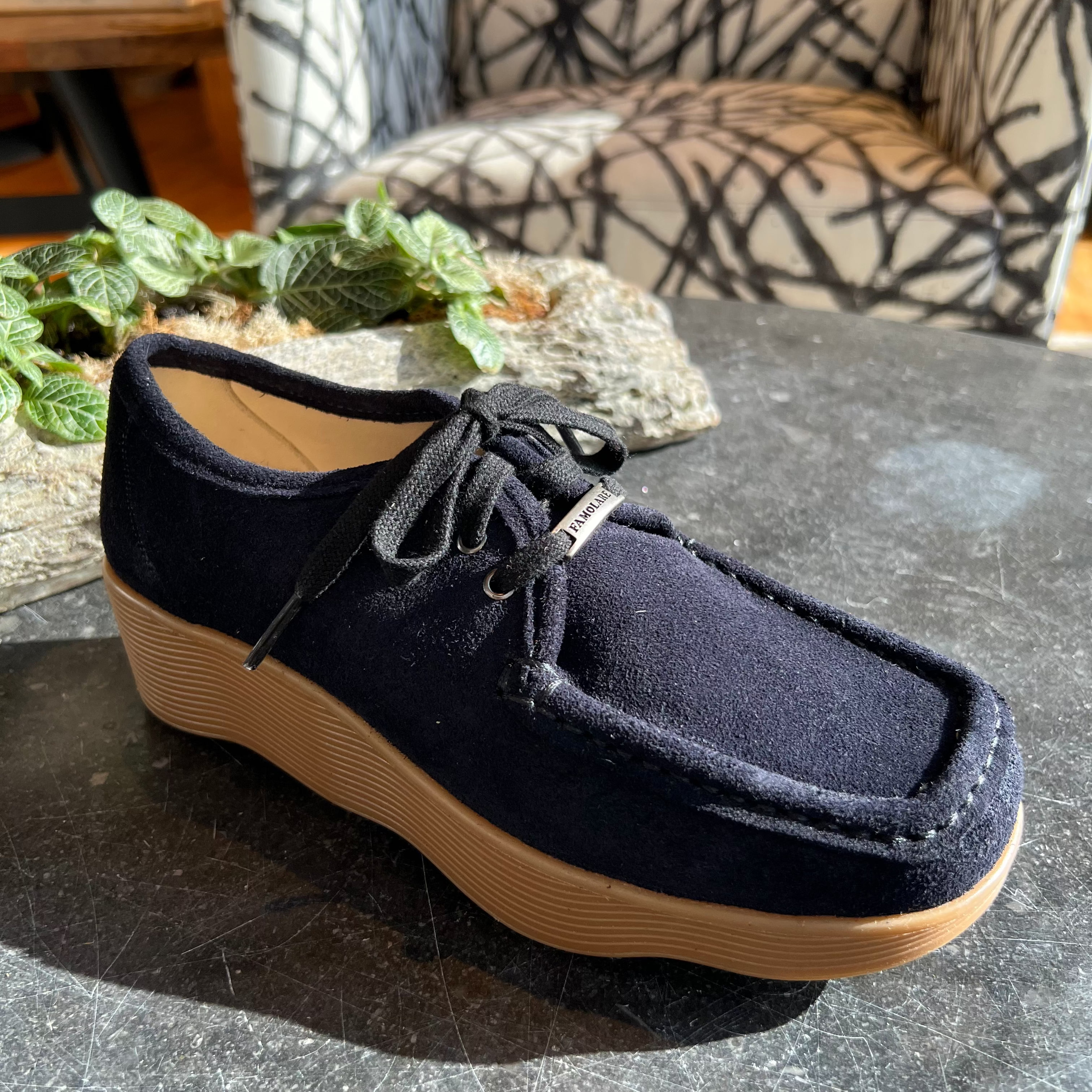 A single navy blue suede slip-on wedge shoe with black laces and a tan sole, resting on a dark stone table in a living room setting.