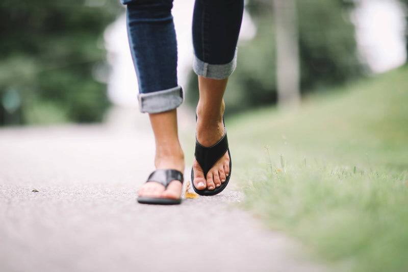 Person wearing black Famolare sandals with rolled denim while walking along a garden path surrounded by soft greenery.