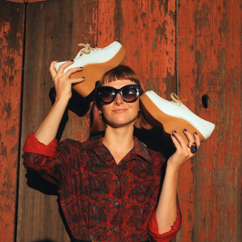 Woman holding white Famolare platform sandals while wearing sunglasses beside a rustic wooden wall.