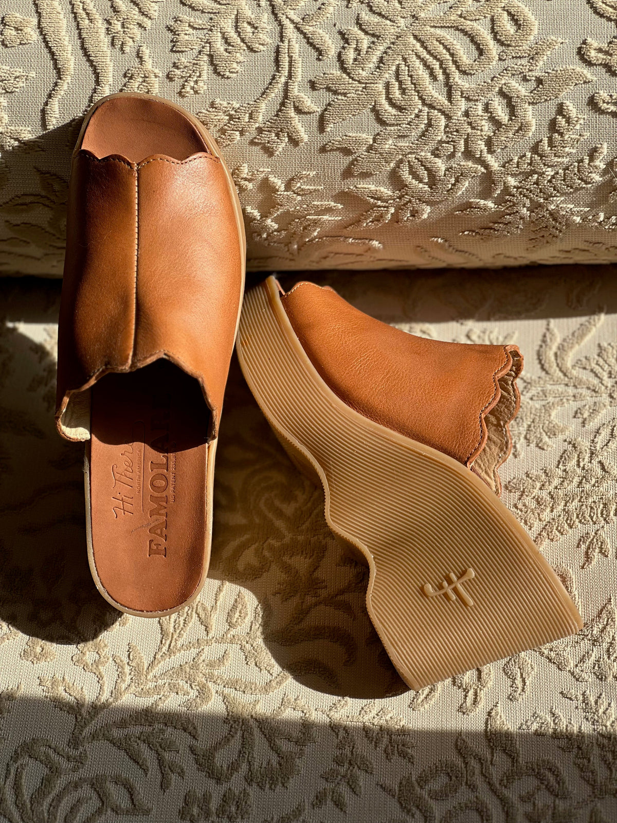 Tan Famolare leather sandals with signature recycled rubber wavy soles resting on a patterned cushion in sunlight.