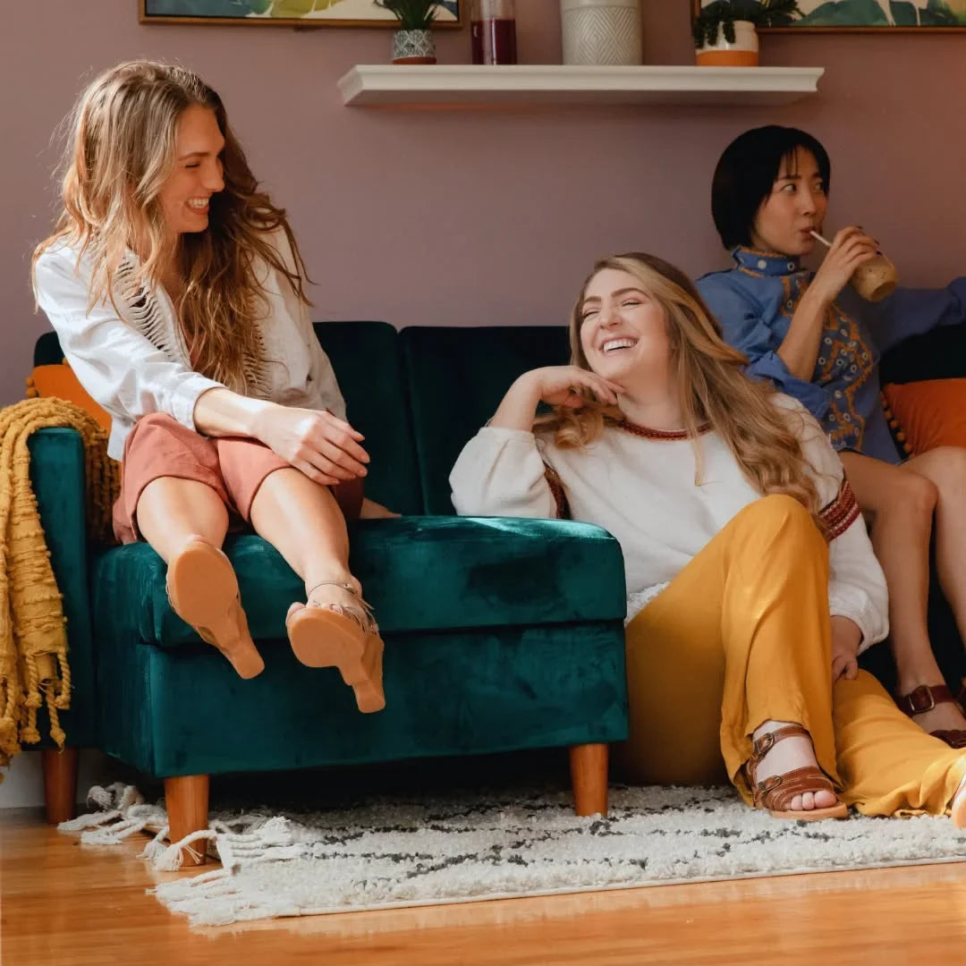 Three women relaxing around a green velvet sofa while wearing retro Famolare shoes in a colorful living room.