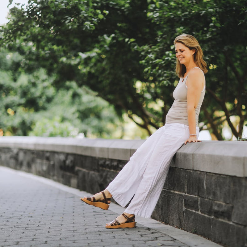 Woman wearing a breezy white linen outfit paired with Famolare sandals while sitting outdoors in a relaxed summer setting.