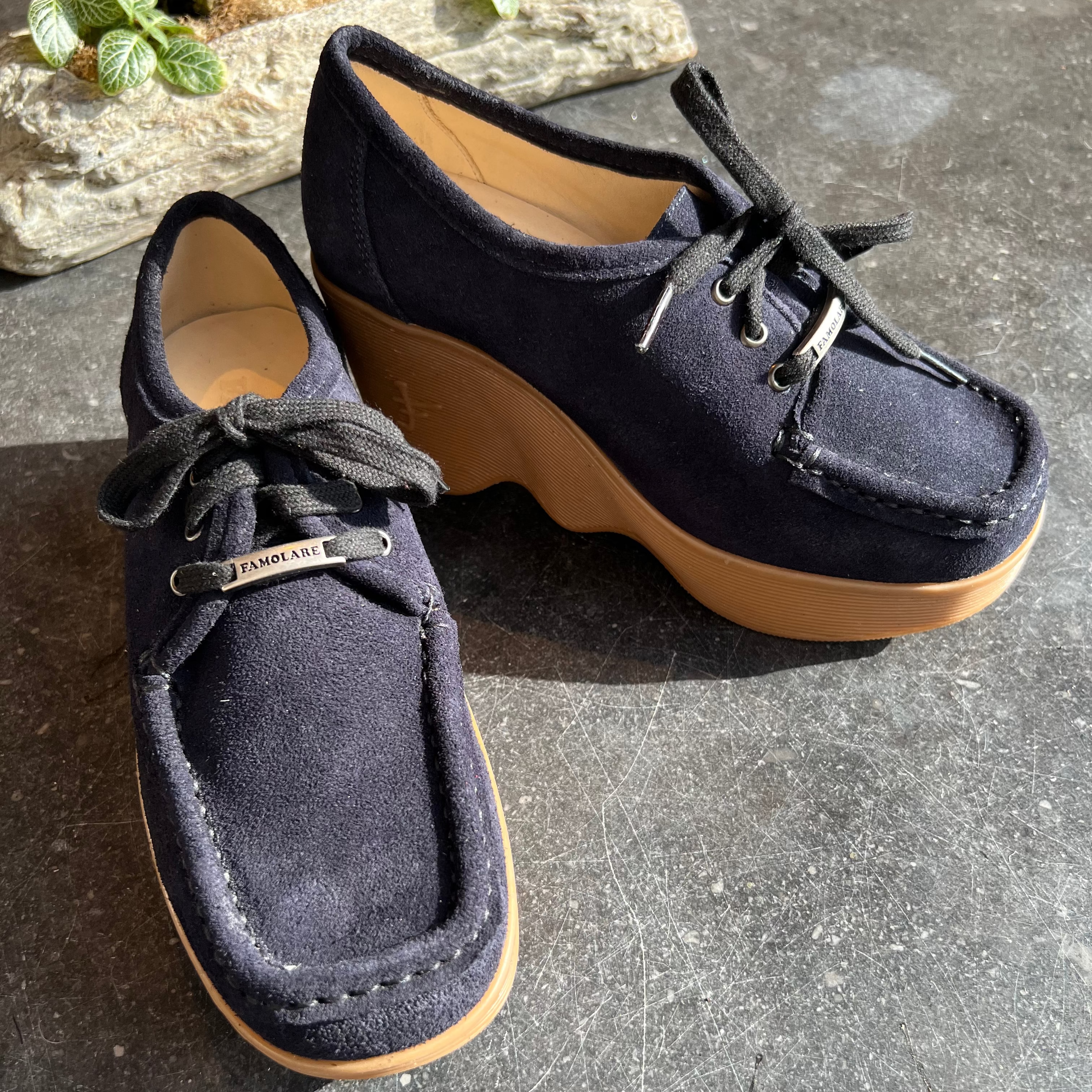 A pair of navy blue suede platform shoes with black laces and a tan rubber sole, displayed on a dark stone surface.